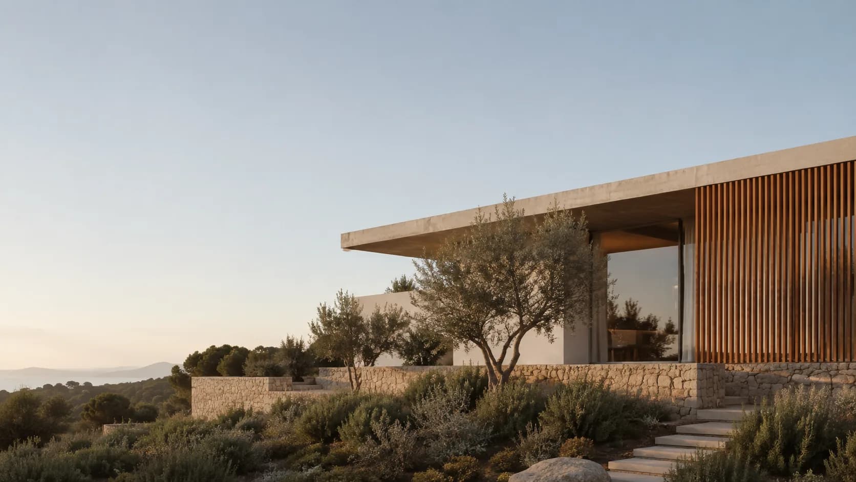 Vivienda de una planta con voladizo de hormigón y celosía de madera, sobre un muro de piedra entre olivos, al atardecer